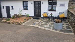 En vit byggnad med ett tak täckt av tegelpannor, en svart dörr och två fönster. En stensatt gång leder till en sittplats med två stolar och ett bord, där varje stol och bord har en gul kudde. En krukväxt med en röd blomma står på bordet.