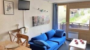 Ein gemütliches Zimmer mit einem blauen Sofa, einem Esstisch mit Stühlen, einem an der Wand befestigten Fernseher und einem Fenster mit Blick ins Freie.