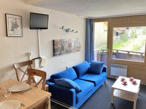 A cozy room with a blue sofa, a dining table with chairs, a TV mounted on the wall, and a window with a view of the outdoors.