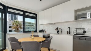 Una cocina moderna con armarios blancos, una mesa de comedor de madera con cuatro sillas y un ventanal con vistas a edificios y una palmera.