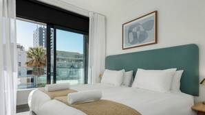 Habitación de hotel con una cama grande, ropa de cama blanca, cabecera color verde azulado y vistas a edificios a través de un ventanal amplio.