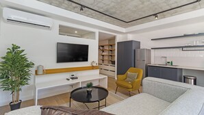 A modern living room with a flat-screen TV, a sofa, a coffee table, and a yellow armchair. There is a kitchen area with a sink and cabinets.