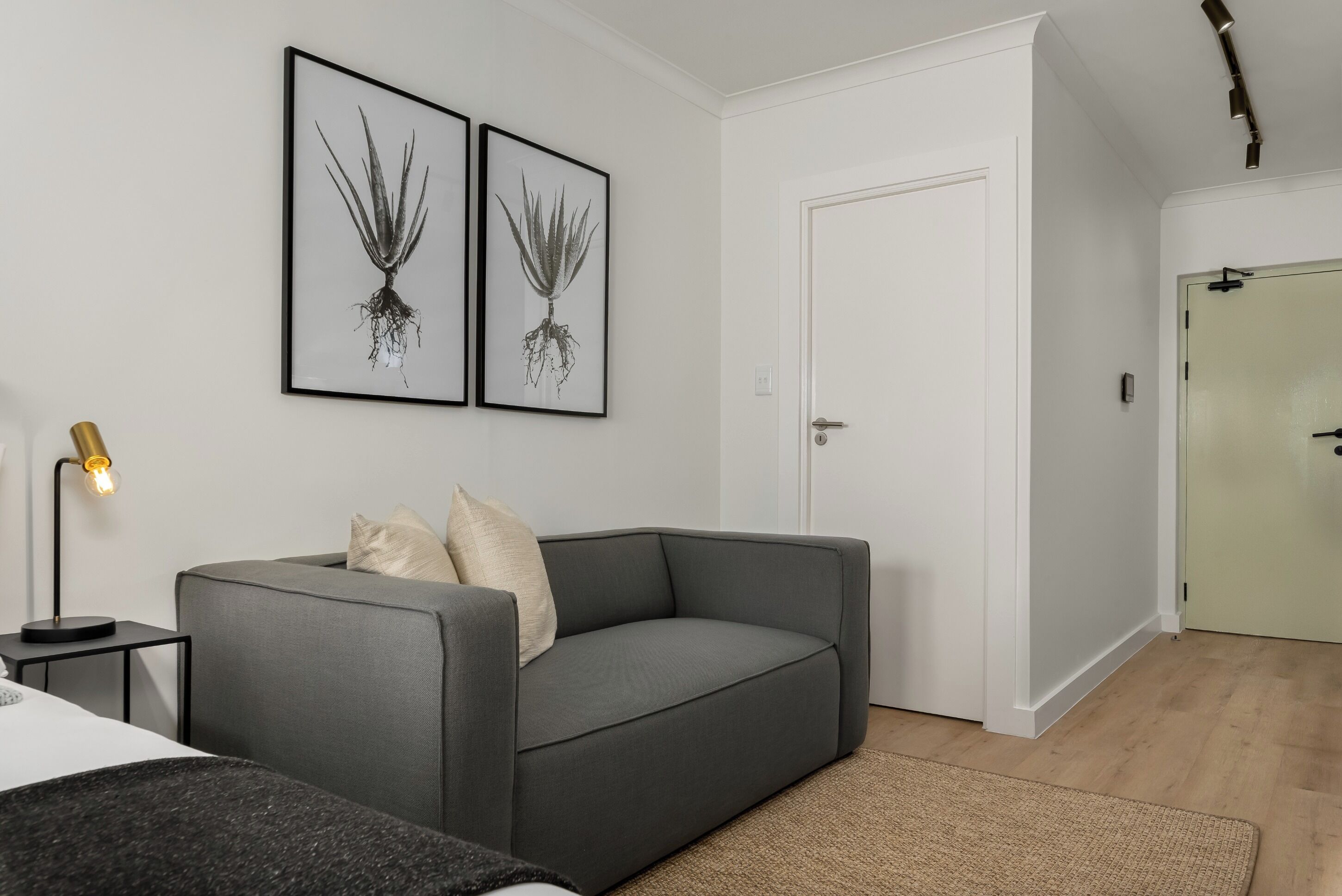 A modern living room with a grey sofa, a small side table with a lamp, and two framed botanical prints on the wall.