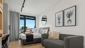 A modern bedroom with a bed, grey sofa, and a desk. There are two framed pictures on the wall, a large window, and a view of buildings outside. - The Sage l Elegant Studio in Vibrant Sea Point (Cape Town)