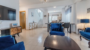 Uma sala de estar espaçosa com uma mesa de centro de madeira escura, um sofá azul e um candelabro. Cozinha visível ao fundo, com uma área de refeições.