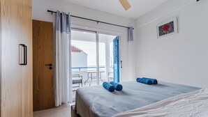Quarto de hotel com uma cama, porta de madeira, varanda com mesa e cadeiras e um quadro na parede.
