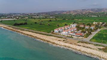 An aerial view of a coastal area with a row of white buildings with red roofs, a stone sea wall, and green fields.