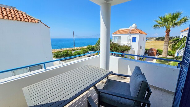 Varanda com mesa e cadeira, vista para área costeira com visão clara do mar e um edifício branco com telhado vermelho.