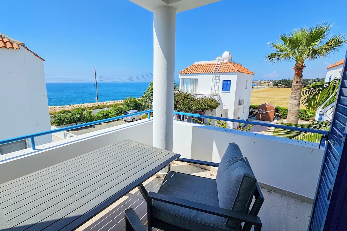 A balcony with a table and chair, overlooking a coastal area with a clear view of the sea and a white building with a red roof.