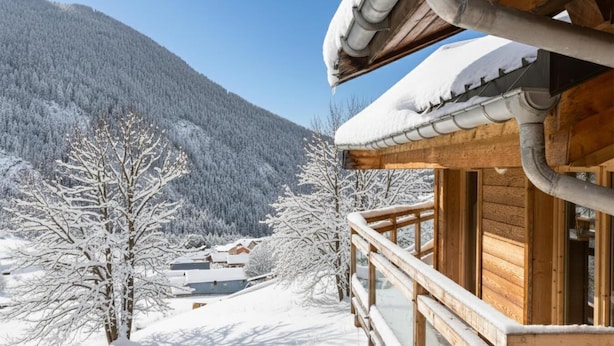 Una cabana de fusta amb un balcó, envoltada d'arbres coberts de neu i una muntanya al fons.