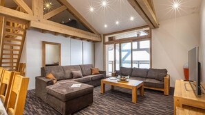 Een ruime woonkamer met een grote hoekbank, een salontafel en een televisie. De kamer heeft zichtbare houten balken, grote ramen en uitzicht op de omgeving.