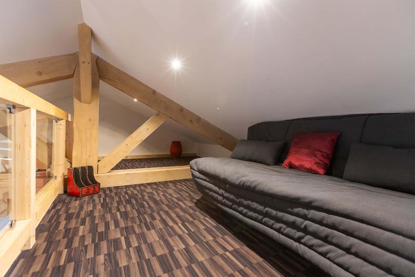 Ein Schlafzimmer im Loft-Stil mit schrägem Dach, Holzbalken, einem grauen Sofa und einem gemusterten Boden.