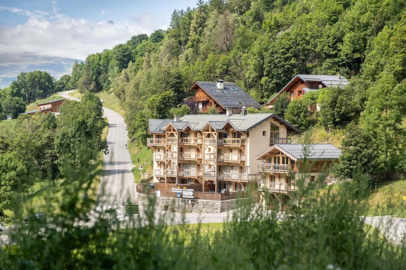 A multi-story lodge with balconies, surrounded by lush greenery and a winding road.
