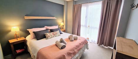 Een net opgemaakt bed met kussens en dekbed, twee nachtlampjes en een raam met gordijnen.