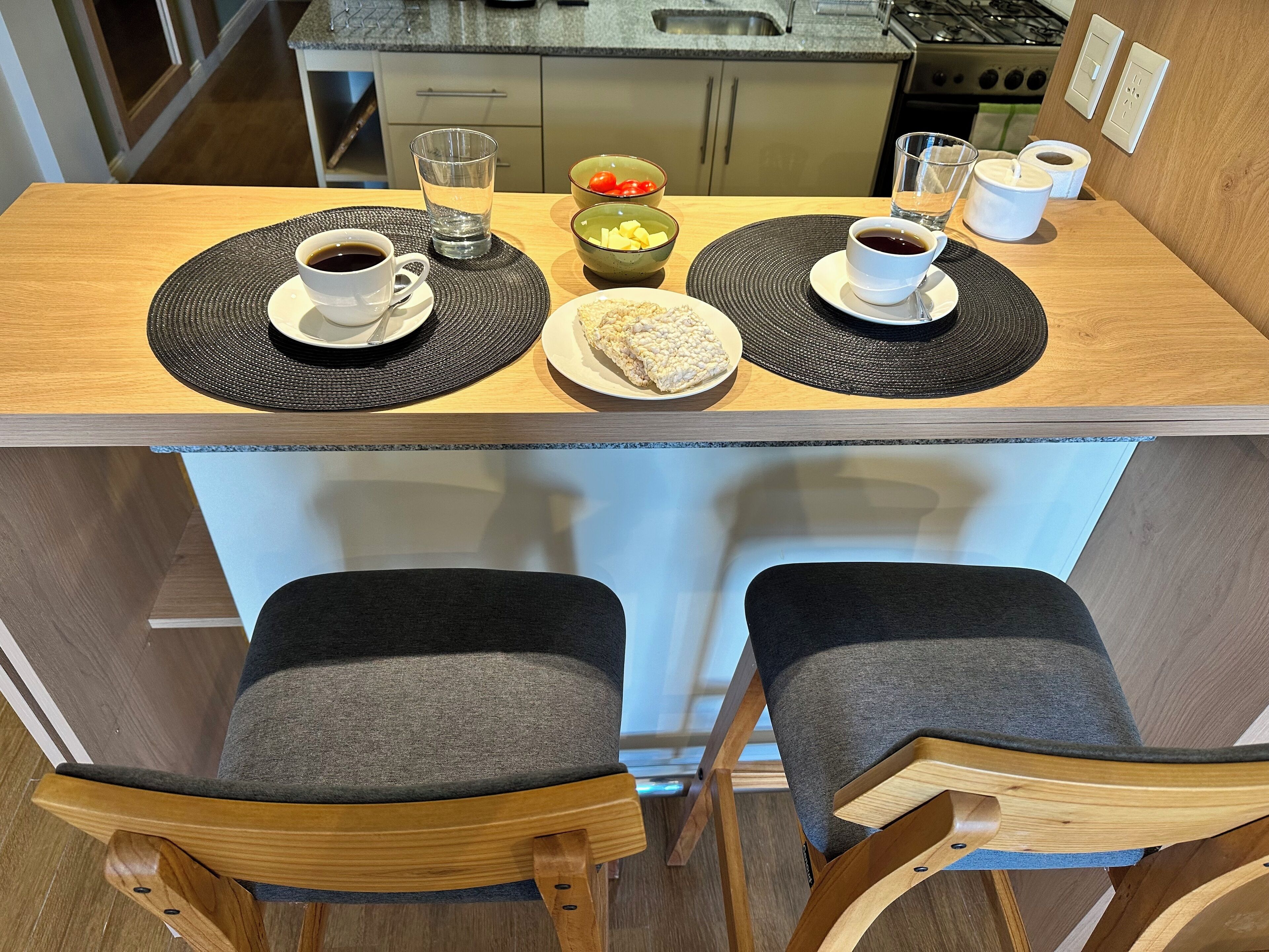 A compact kitchen with a small dining area, including a counter with two chairs and a table set with a bowl of fruit, a plate of rice, and two cups of coffee.