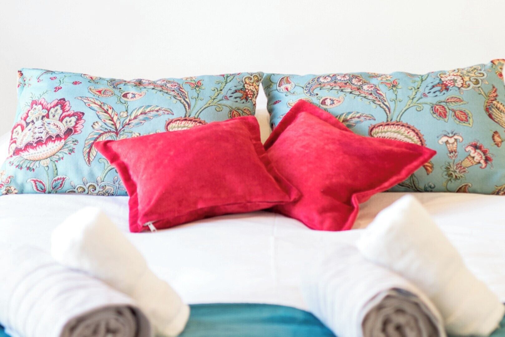 Una cama con un cojín estampado con flores y dos cojines rojos, ropa de cama blanca y calcetines grises y blancos enrollados.