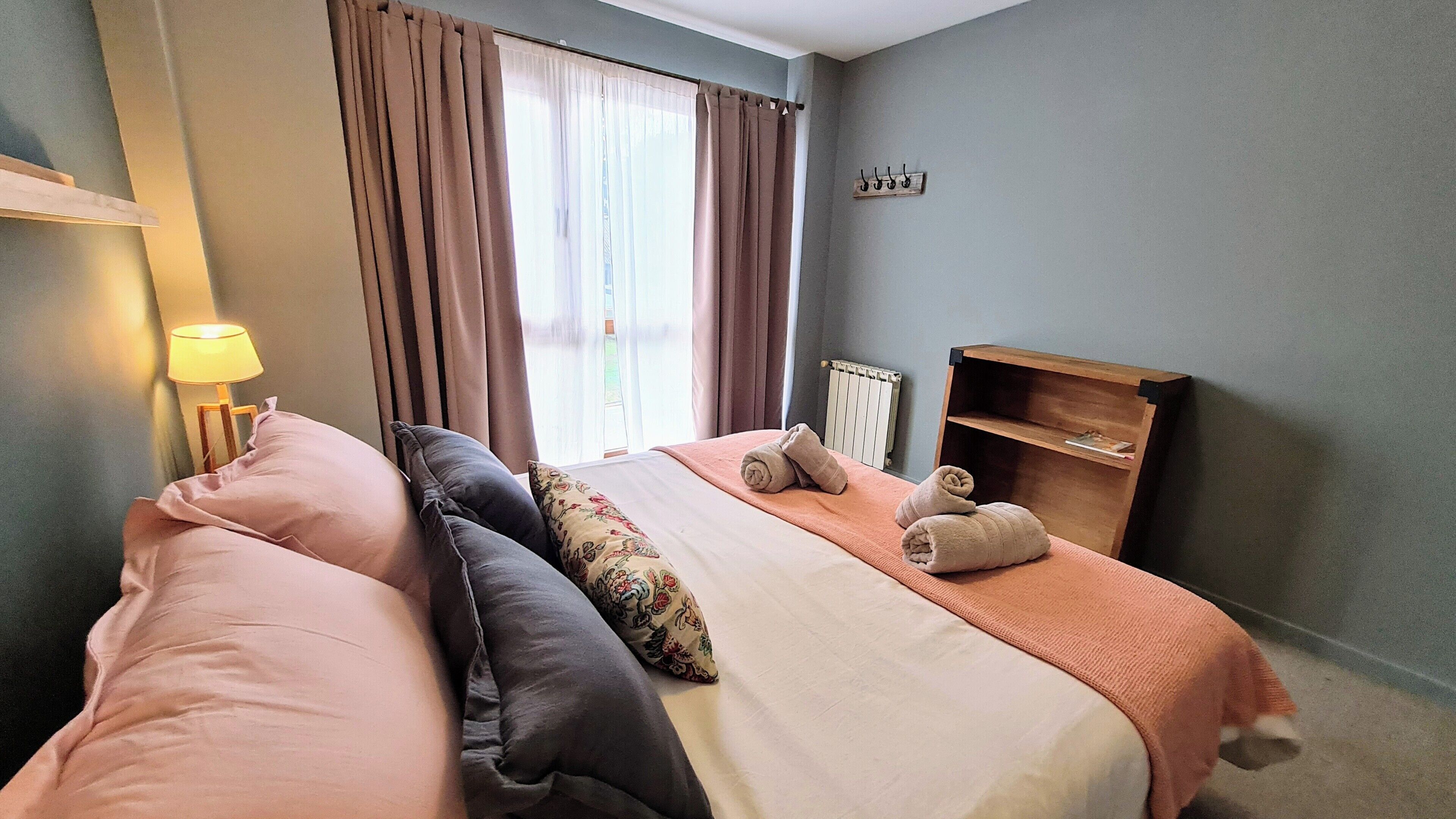 Habitación de hotel con cama, almohadas, mesita de noche, lámpara, ventana con cortinas y radiador.