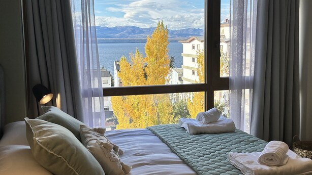 A hotel room with a bed, pillows, a quilted blanket, and a view of a lake and mountains through a window. - Terrazas I y II (San Carlos de Bariloche)