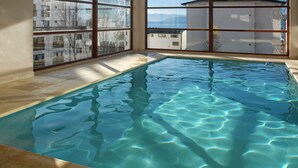 An indoor swimming pool with clear water, large windows, and a view of buildings outside. - Terrazas I y II (San Carlos de Bariloche)