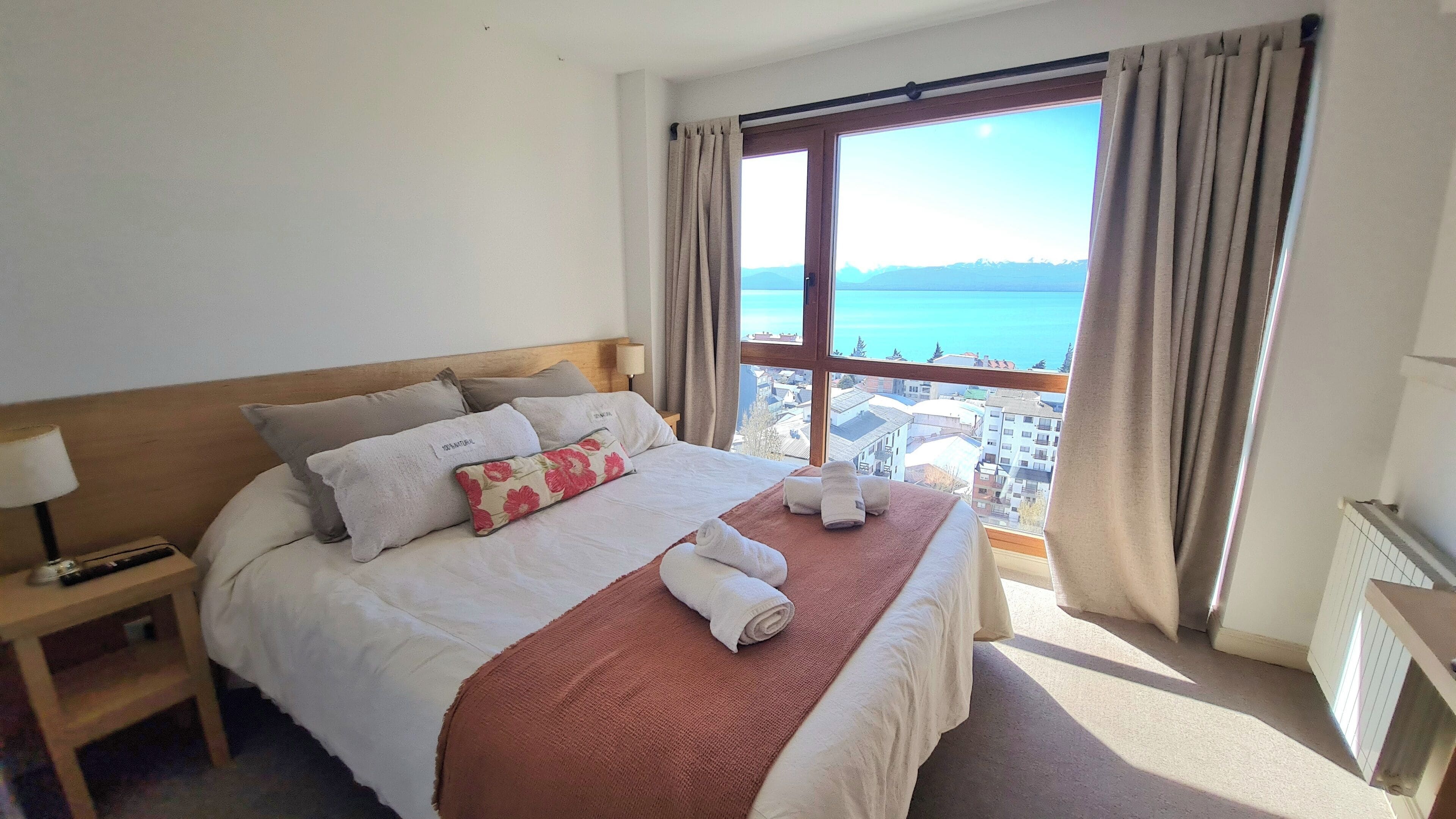 A hotel room with a large bed, a nightstand, a window with a view of the city, and a view of the sea.