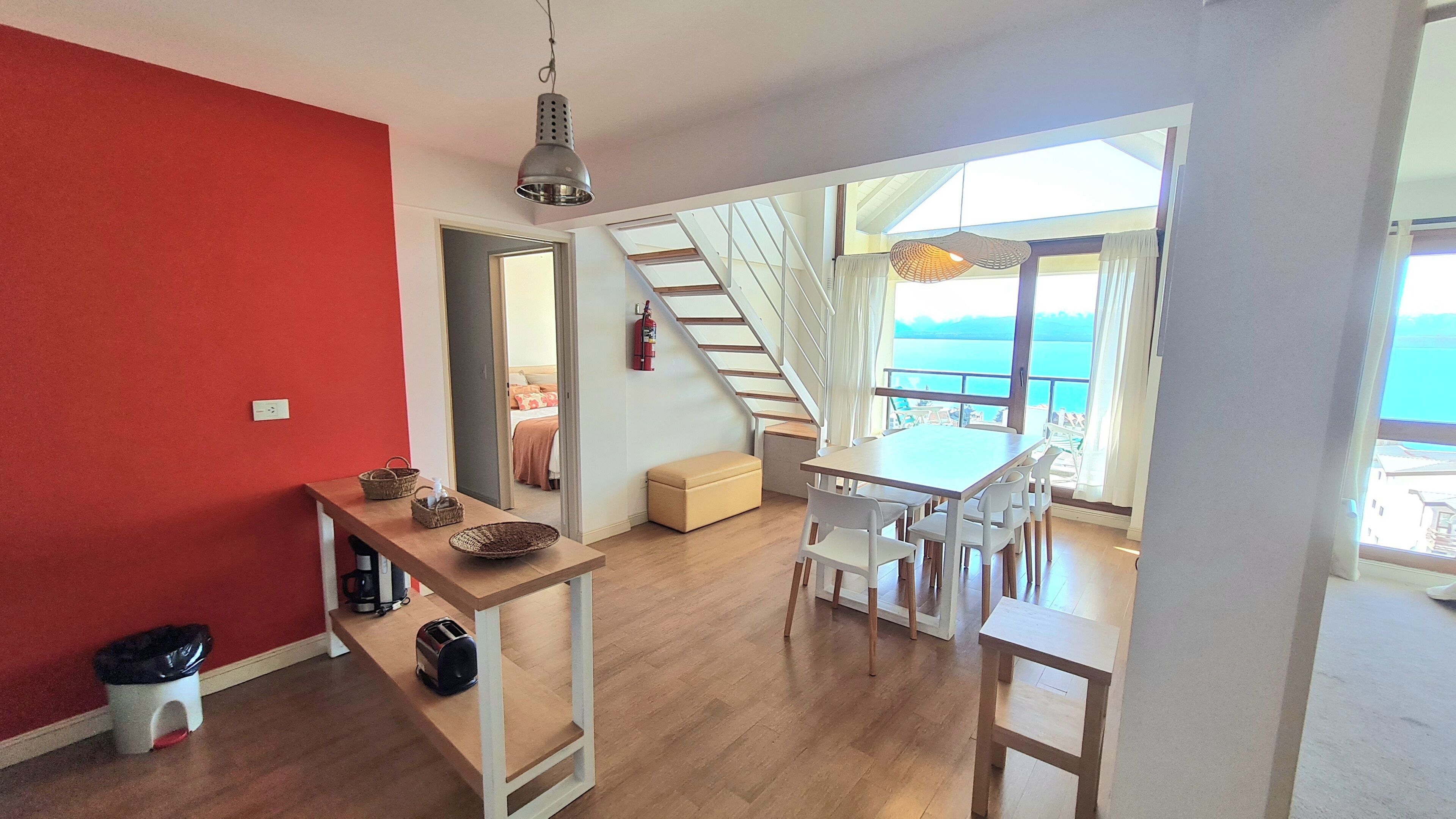 Une cuisine moderne avec un plancher en bois, un coin repas avec des chaises blanches, un escalier menant à un balcon avec vue sur la mer et un mur d’accent rouge.