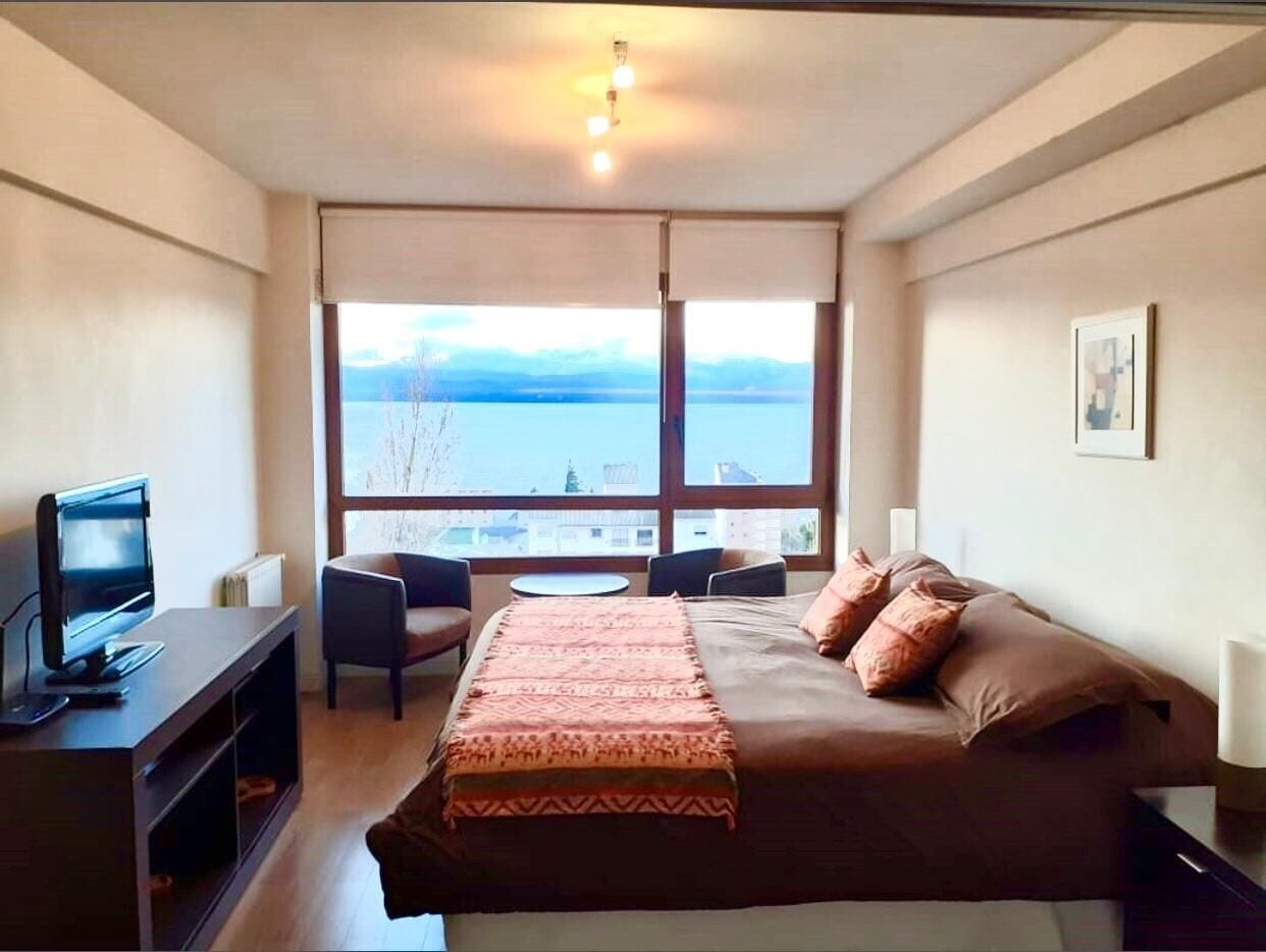 Habitación de hotel con cama, televisor, escritorio y silla. Amplia ventana con vista a un cuerpo de agua y montañas.