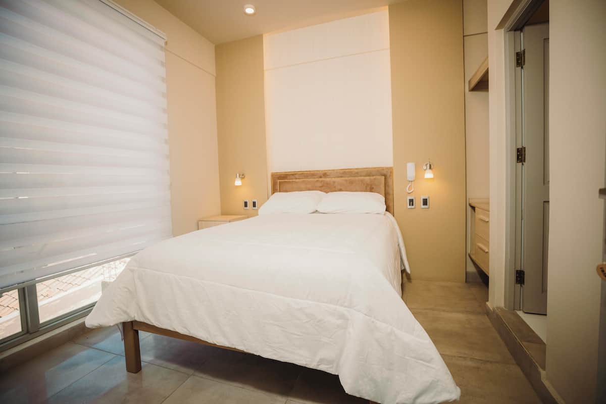 A bedroom with a bed, bedside lamps, a window with blinds, and a door.