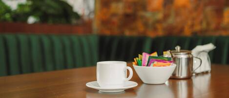 A wooden table with a white cup and saucer, a bowl of colorful pencils, and a silver coffee pot.