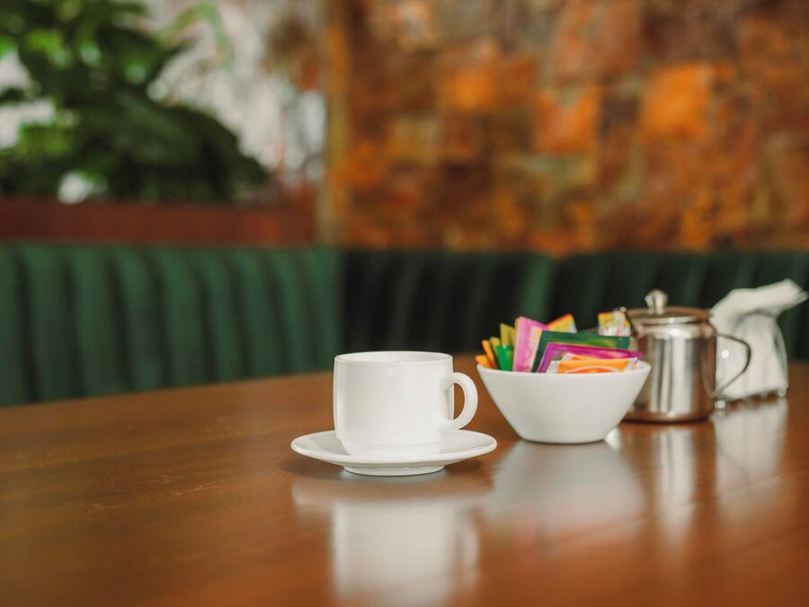 Een houten tafel met een witte kop en schotel, een schaal vol kleurrijke potloden, en een zilveren koffiepot.