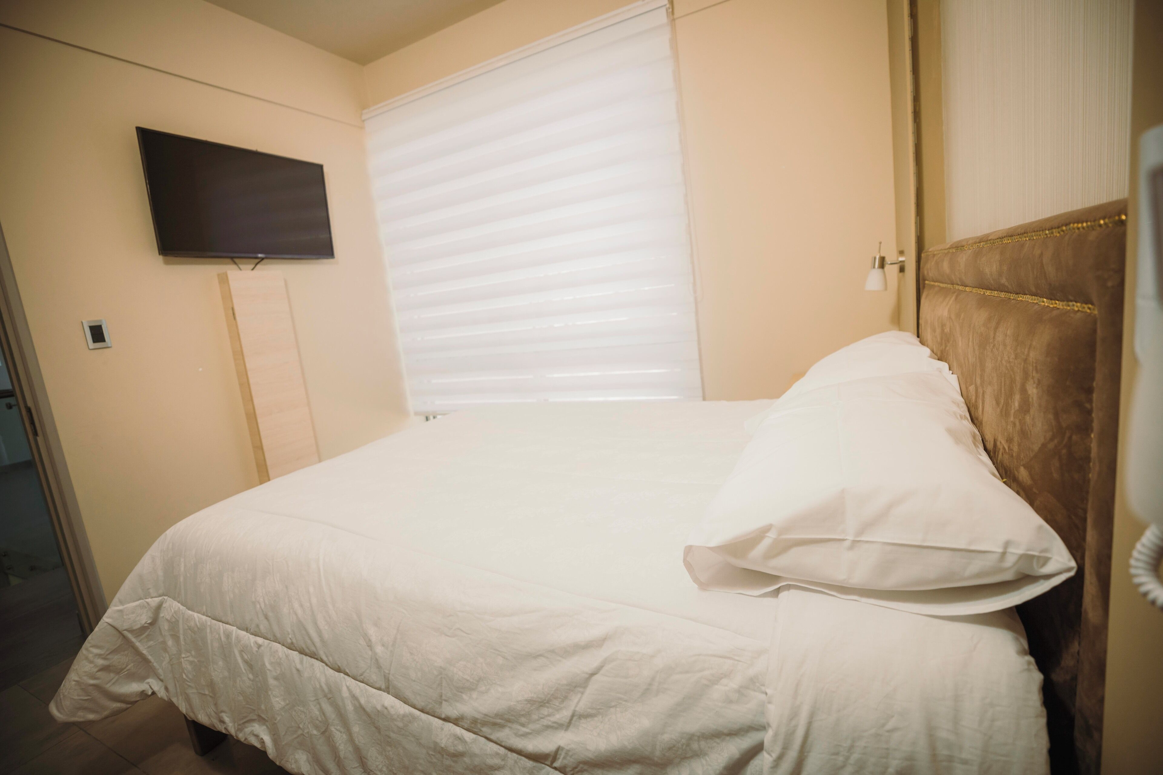 A hotel room with a bed, a television mounted on the wall, and a window with blinds.