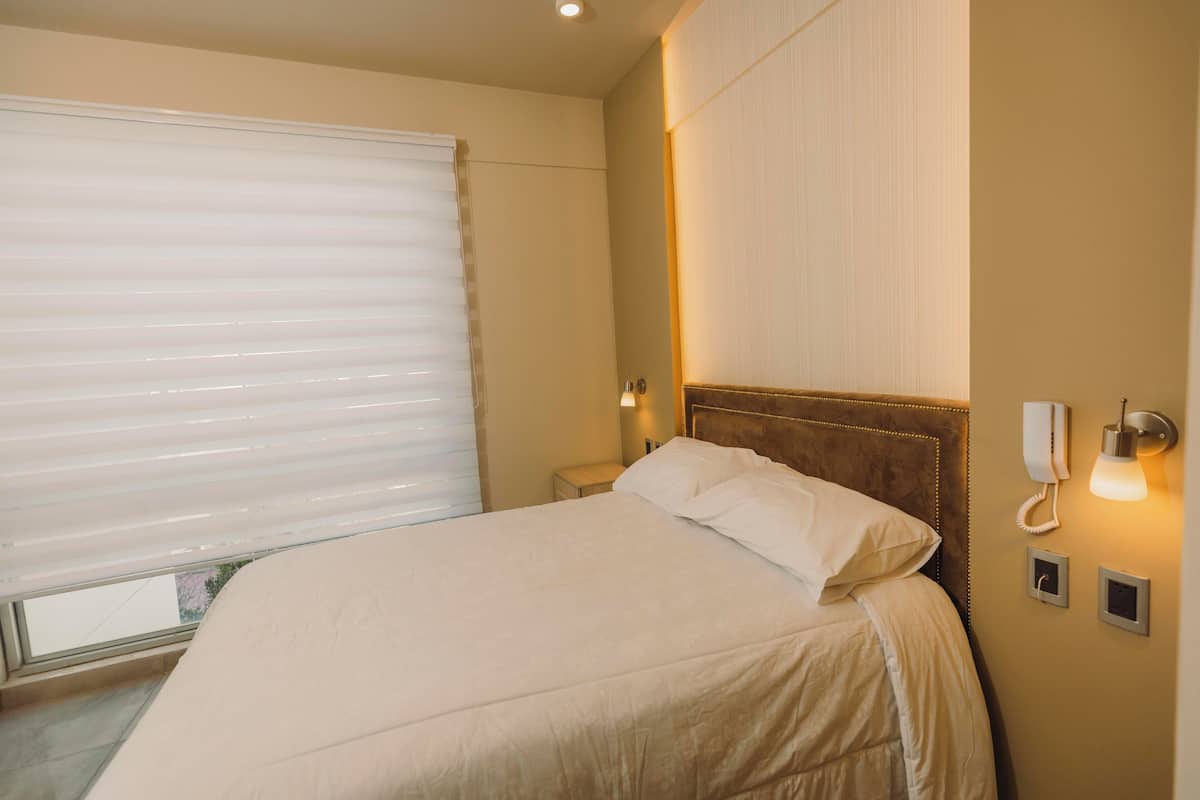 A hotel room with a large bed, a bedside table, a lamp, a phone, and a window with blinds.