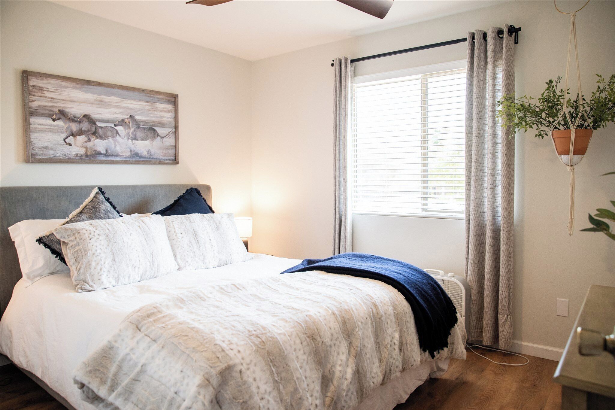 Un dormitorio con cama, ventilador de techo, ventana con cortinas, una planta en la pared y un cuadro.
