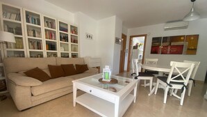 Un salon avec un canapé beige, une table basse en verre, des chaises blanches et une bibliothèque remplie de livres.