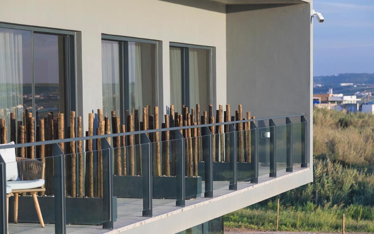 Un edificio moderno con grandes ventanales, un balcón con pilares de madera y una silla.