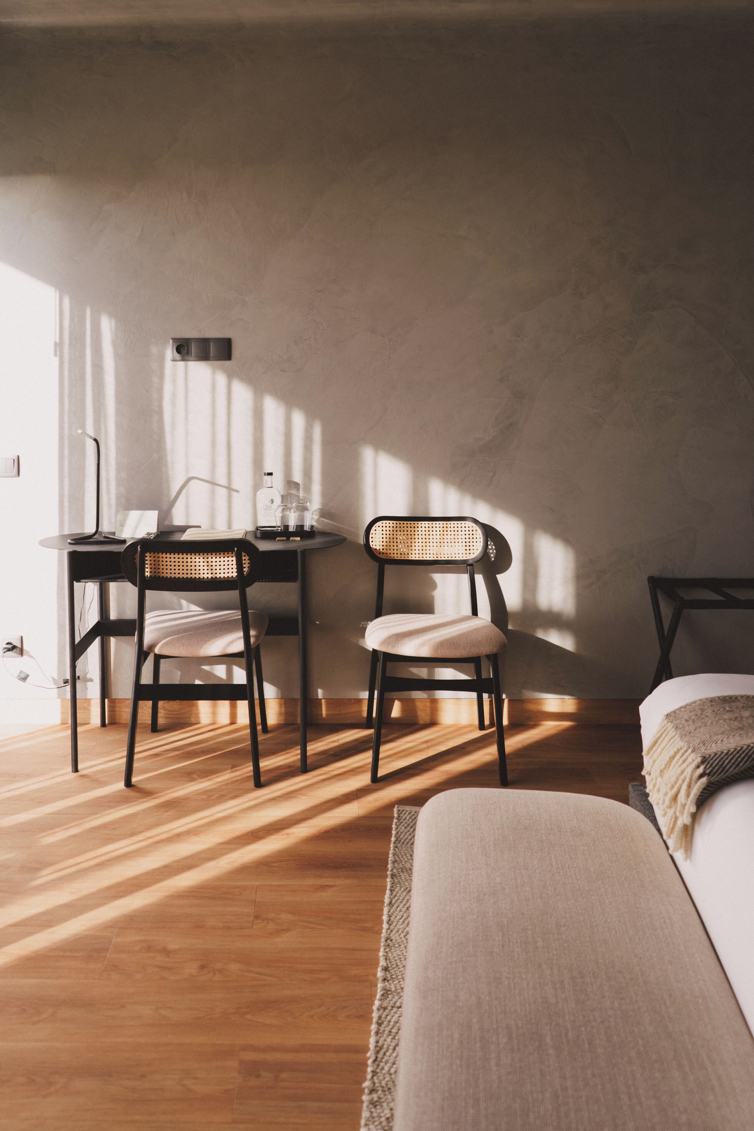 Una habitación con piso de madera, dos sillas, una mesita, una lámpara y una cama con una colcha.