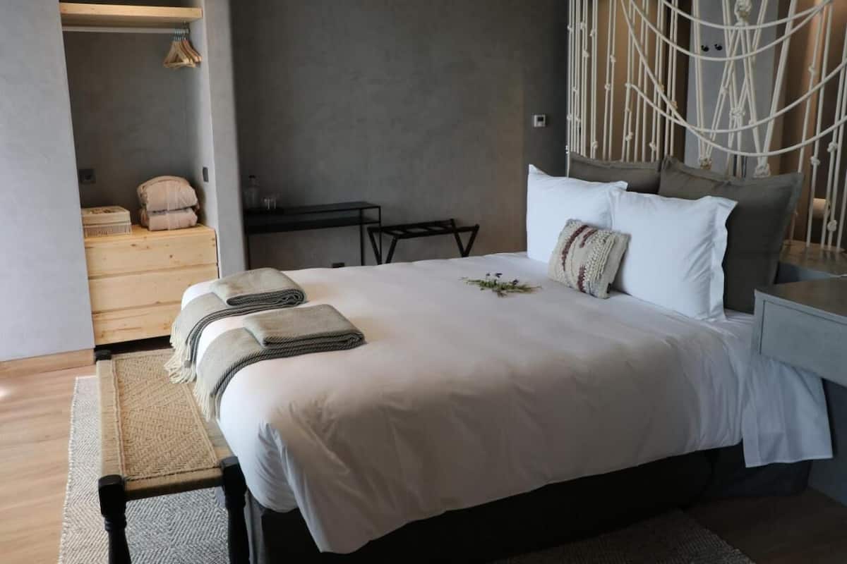 Een net opgemaakt bed met witte lakens, een klein nachtkastje en een macramé wanddecoratie.