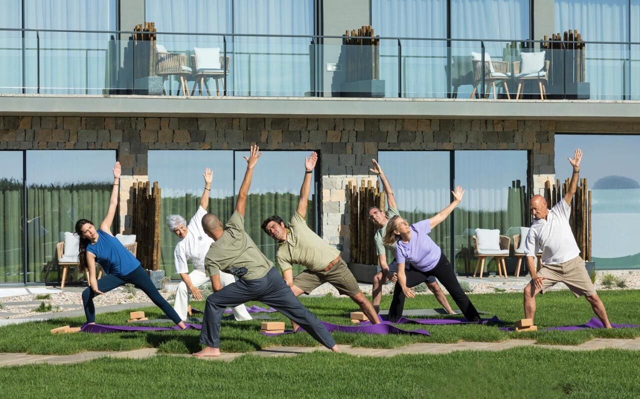 Un groupe de personnes pratiquant des exercices de yoga en plein air, devant un bâtiment moderne doté d’une façade en pierre et de grandes fenêtres.