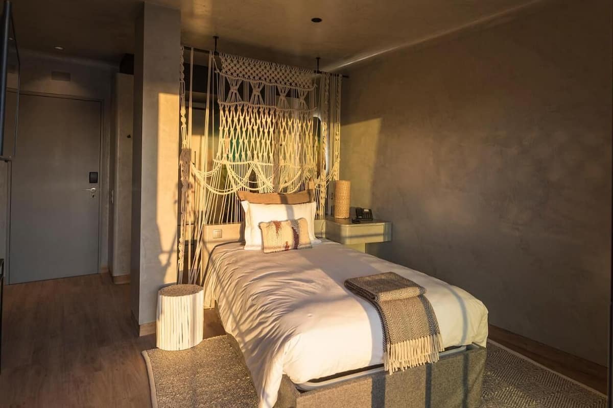 Een slaapkamer met een bed, nachtkastjes, een macramé hoofdbord bevestigd aan de muur, en een klein houten krukje.
