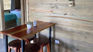 Interior de madera con una mesa, taburetes y una cama al fondo. Un cartel en la pared dice: 'La vida es mejor con buen café'.
