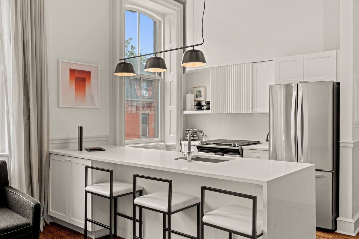 A modern kitchen with a large island, bar stools, a stainless steel refrigerator, and pendant lighting.