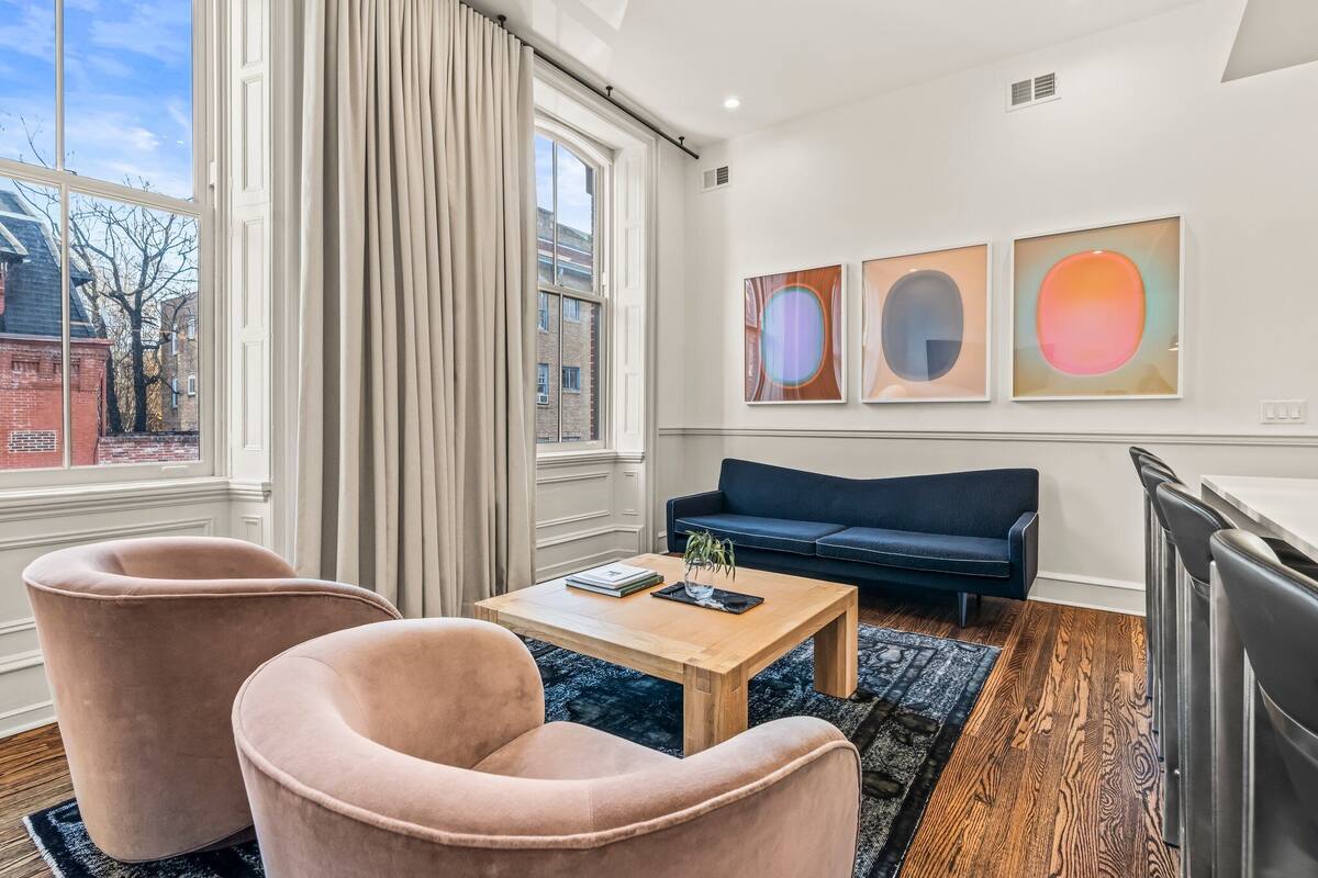 A modern living room with a large window, a dark blue sofa, two pink armchairs, a wooden coffee table, and three abstract paintings on the wall.