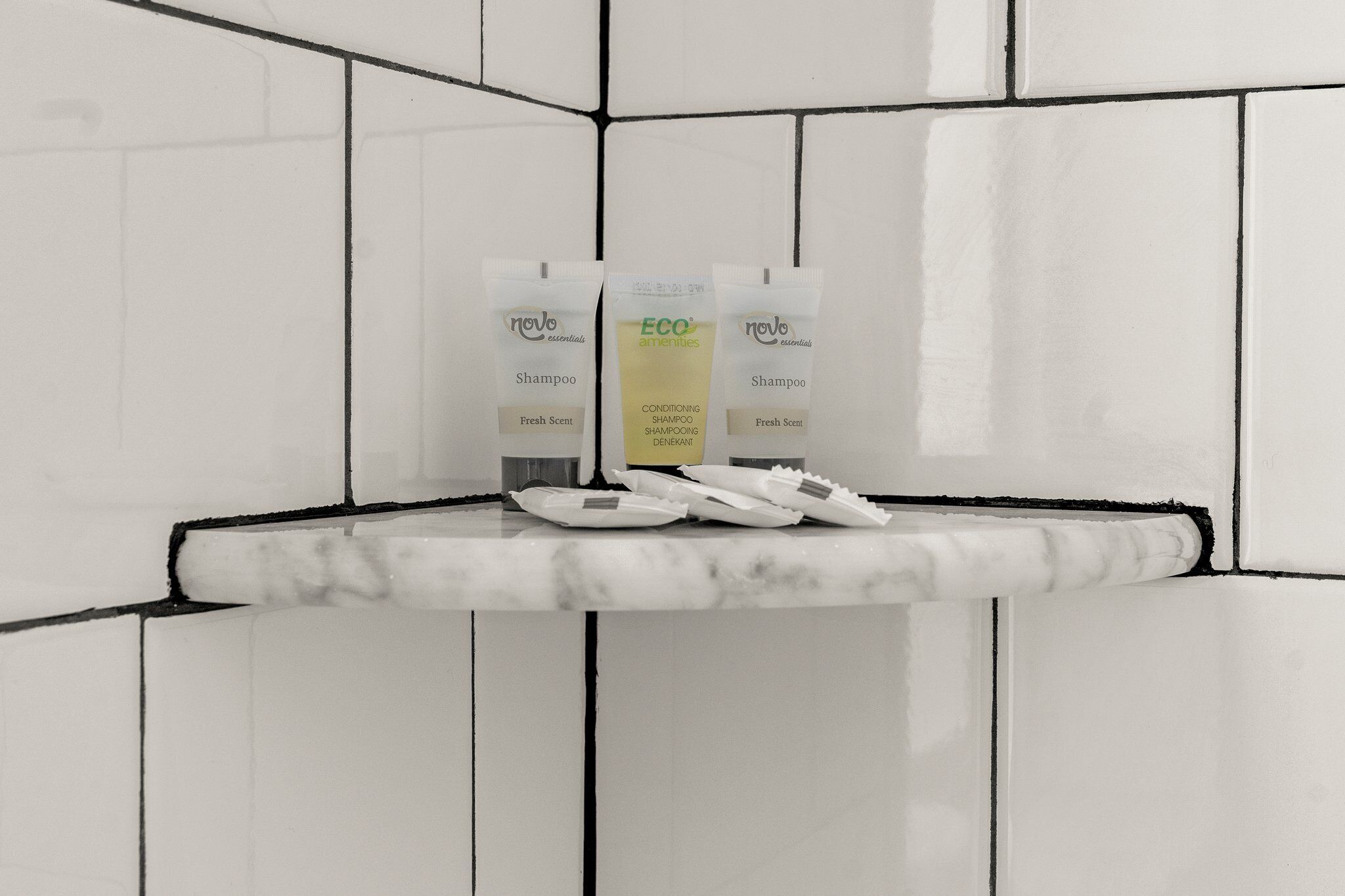 A bathroom corner with white tiles and a marble countertop. Three toiletries are visible: two 'novo essentials' shampoo bottles and one 'eco amenities' conditioner bottle.