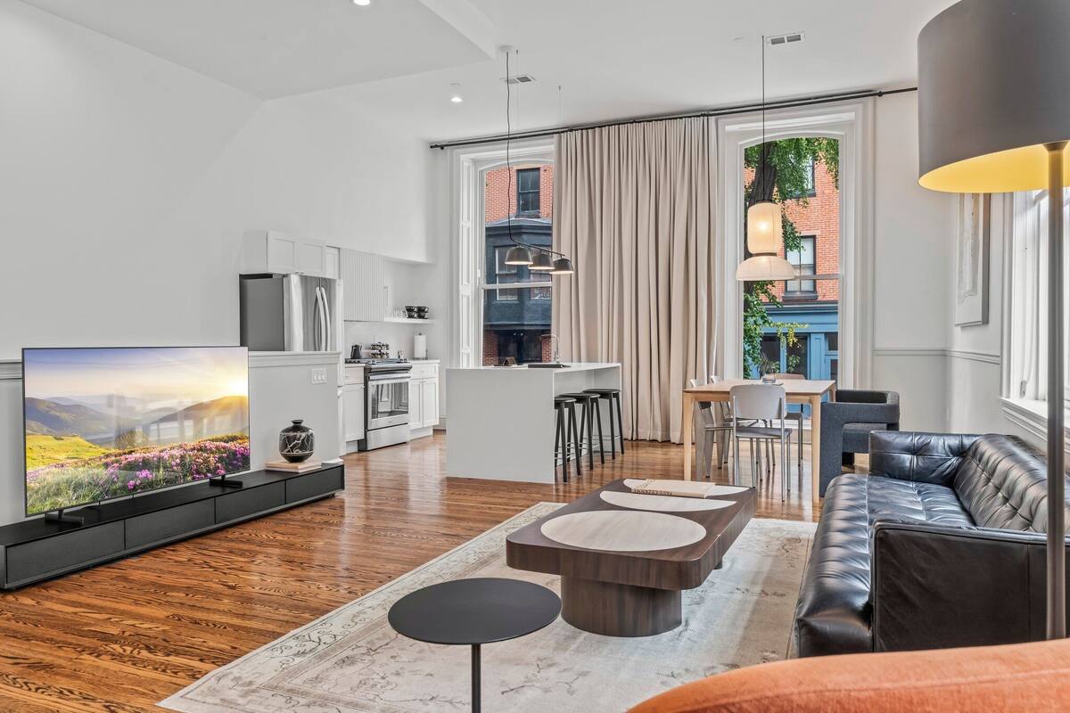 A modern living room with a flat-screen TV, leather sofa, and a dining area with a kitchen in the background.