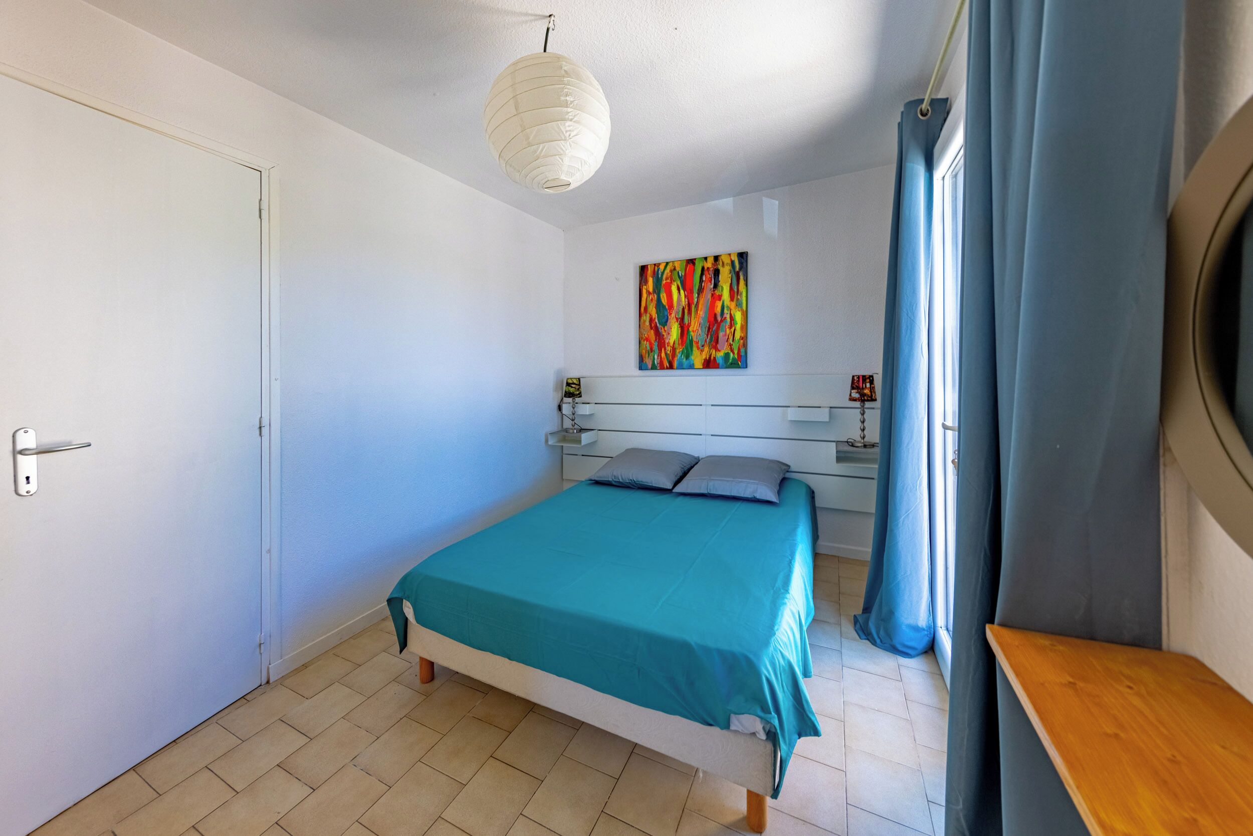 Een slaapkamer met een bed, een nachtkastje, een kleurrijk abstract schilderij, een raam met gordijnen en een houten bankje.