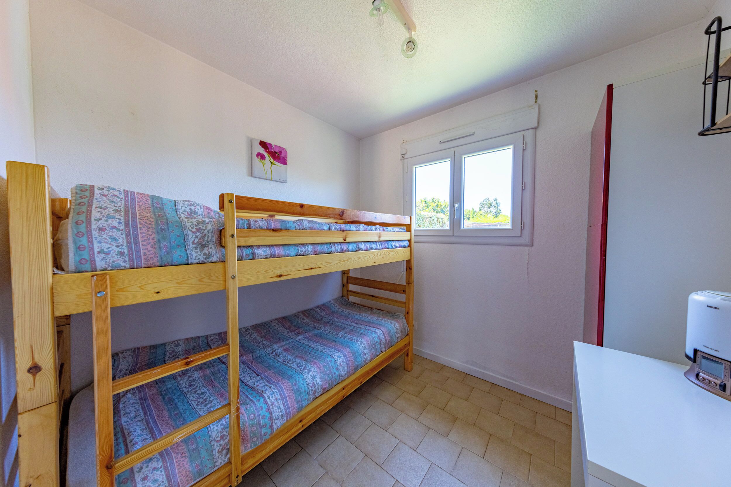 Uma cama beliche com roupa de cama estampada, uma janela com vista e uma escrivaninha branca com um dispositivo.
