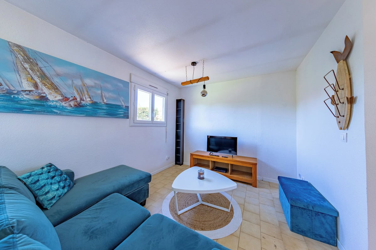 A living room with teal sofas, a white coffee table, a painting of sailboats, a TV on a wooden stand, and a wall-mounted shelf.