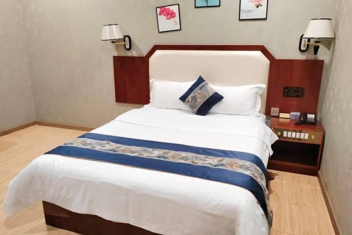 Un lit bien fait, recouvert d’une couvre-lit blanche et bleue, avec une tête de lit en bois et une table de chevet sur laquelle se trouvent une lampe et une horloge.