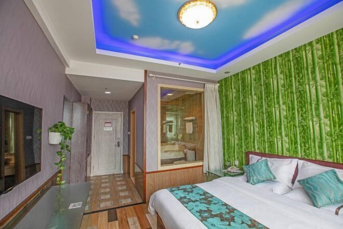 Une chambre d’hôtel avec un lit, une table en verre, un miroir, une porte et une salle de bain visible à travers une paroi en verre.