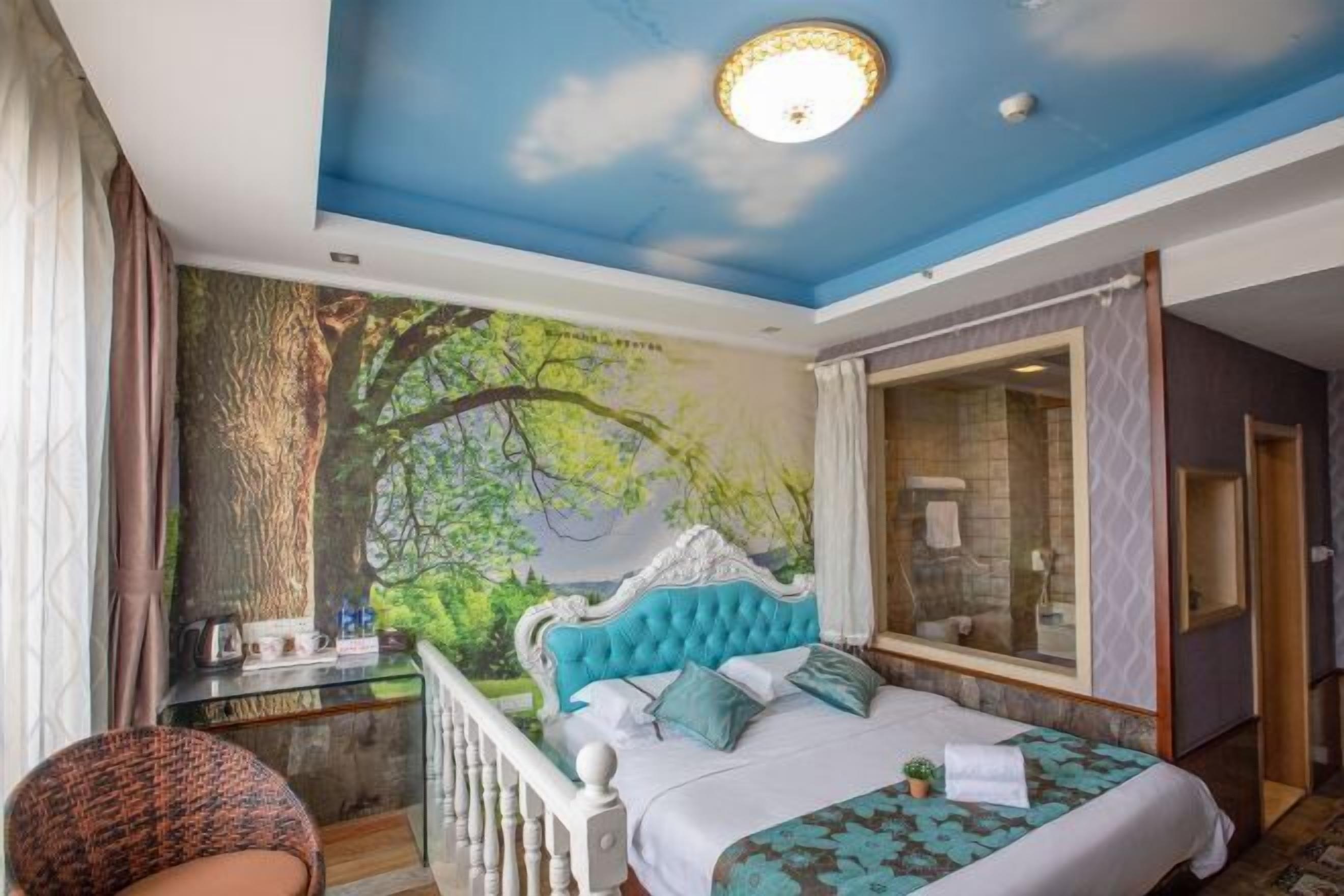 Une chambre à coucher avec une grande fresque représentant une forêt, une tête de lit capitonnée, un lit avec une literie à motifs, un bureau avec une chaise et une salle de bain dotée d’une cabine de douche en verre.