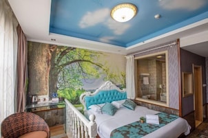 Un dormitorio con un gran mural de un bosque, un cabecero capitonado, una cama con ropa de cama estampada, un escritorio con una silla y un baño con cabina de ducha de vidrio.